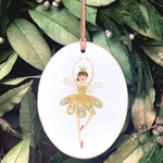 An oval white Christmas ornament featuring a painted ballerina in a yellow tutu with colorful patterned wings, hanging from a brown ribbon against a background of green pine branches and leaves.