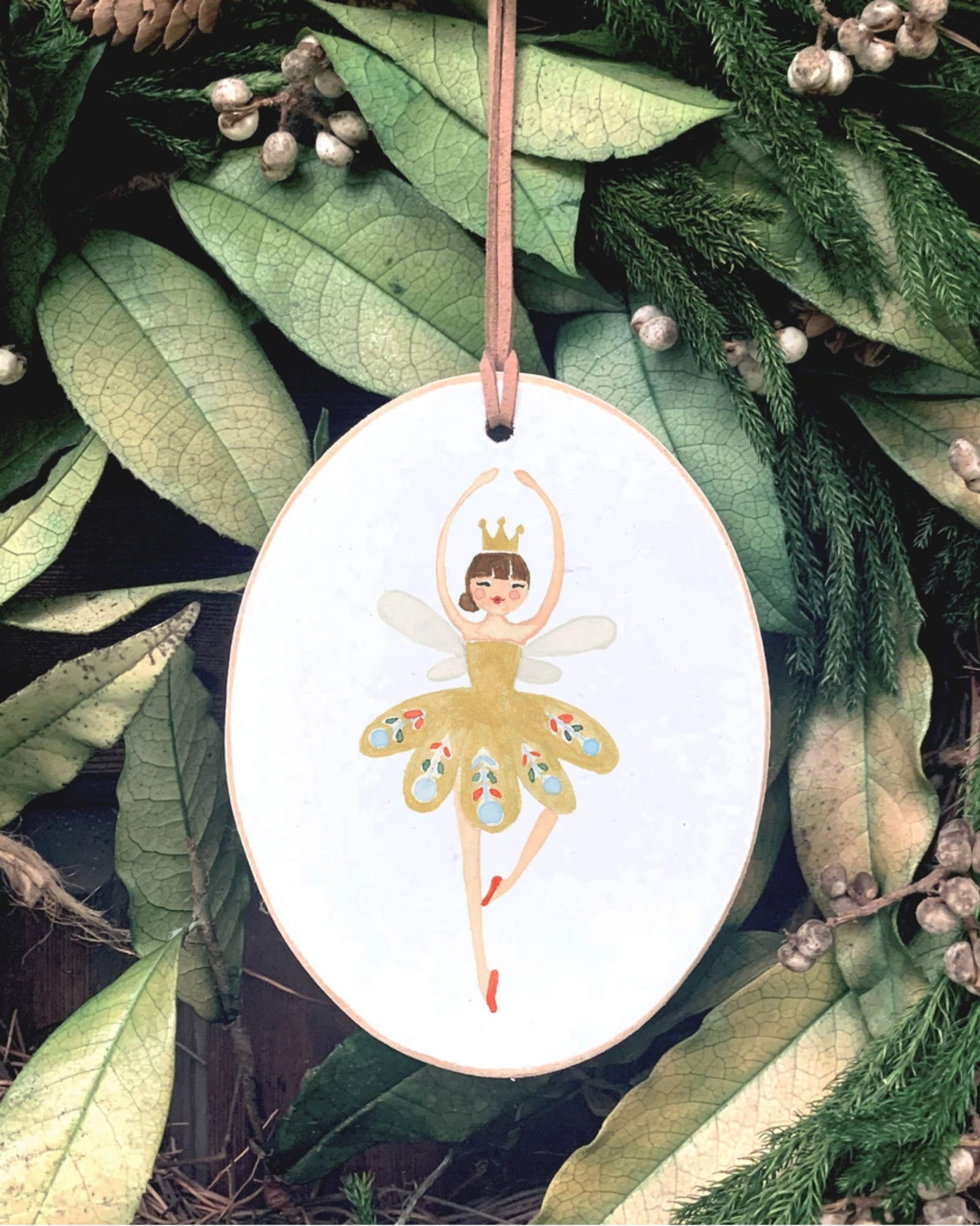 An oval white Christmas ornament featuring a painted ballerina in a yellow tutu with colorful patterned wings, hanging from a brown ribbon against a background of green pine branches and leaves.