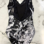 A black and white floral print one-piece leotard with a black lace V-neck bodice, laid flat on a white marble surface.