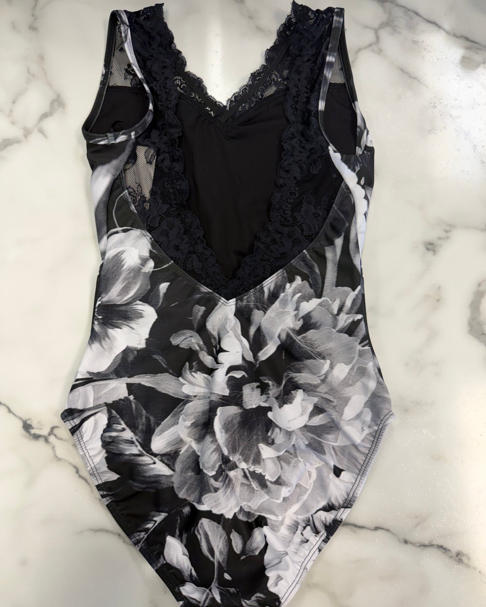 A black and white floral print one-piece leotard with a black lace V-neck bodice, laid flat on a white marble surface.