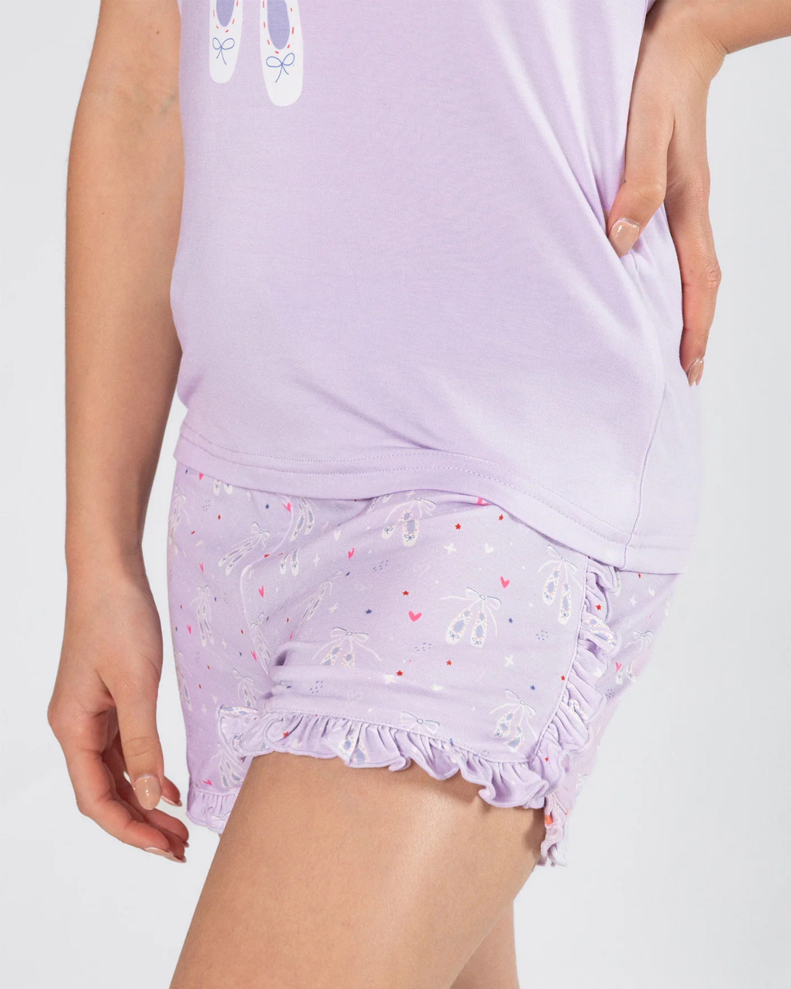 A close-up of a person wearing a light lavender pajama top and matching ruffled shorts with a ballet dancer print and small heart motifs, against a plain light gray background.