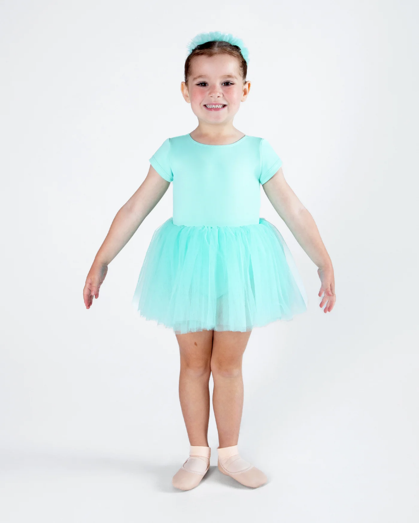 A young girl modeling a light mint green short-sleeve tutu dress with a matching headband and ballet slippers, standing against a plain white studio background.