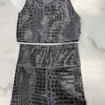 A two-piece activewear set featuring a sleeveless tank top and matching high-waisted shorts, both in a gray and black crocodile-embossed pattern, laid flat on a white marble background.