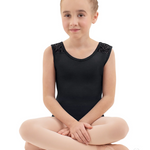 A young girl with blonde hair in a bun sits cross-legged in a black sleeveless ballet leotard with lace-up shoulder detailing, paired with light pink tights and ballet slippers, against a solid white background.