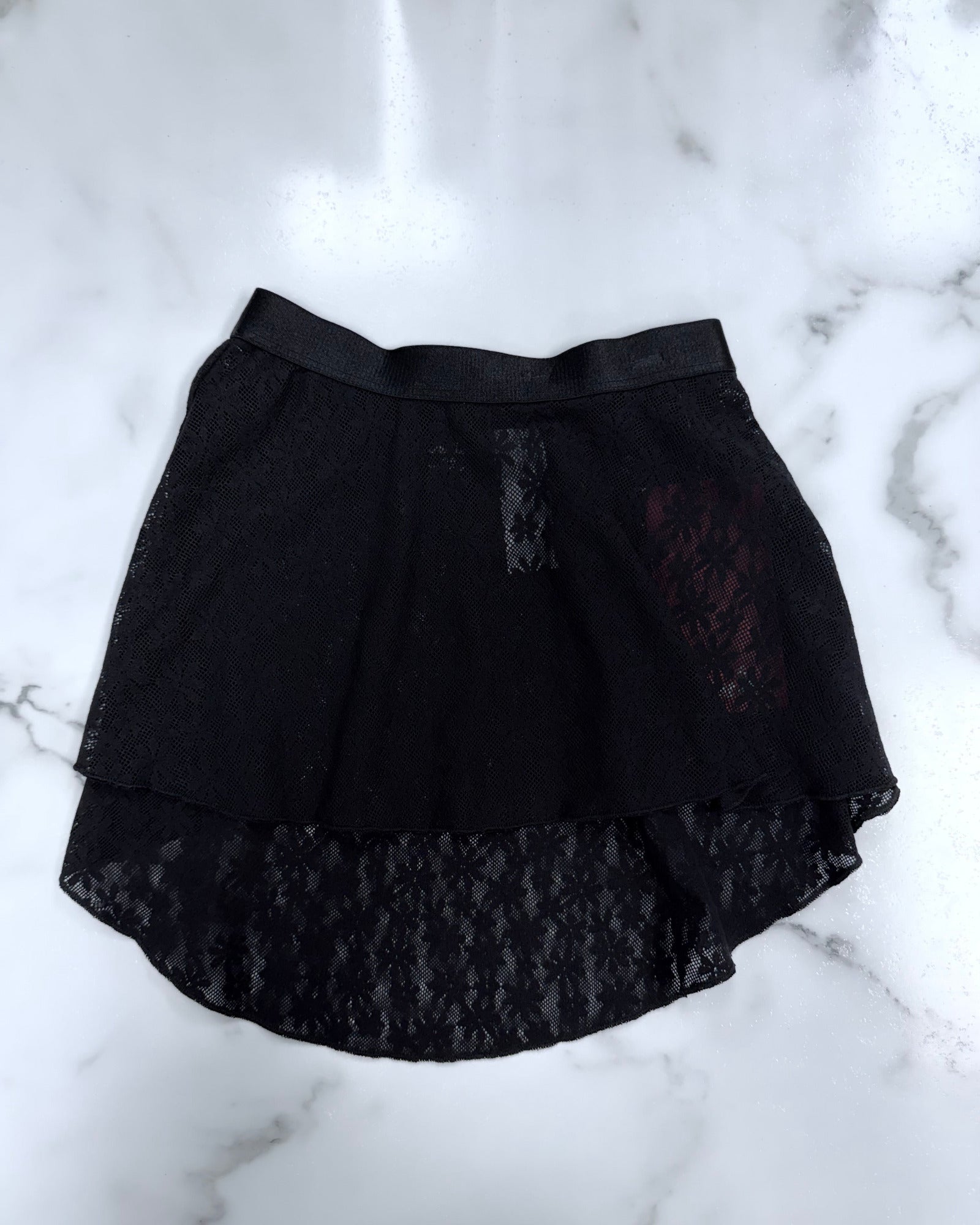 A black lace high-low skirt with a sheer mesh overlay and a solid black waistband, laid flat on a white marble surface.