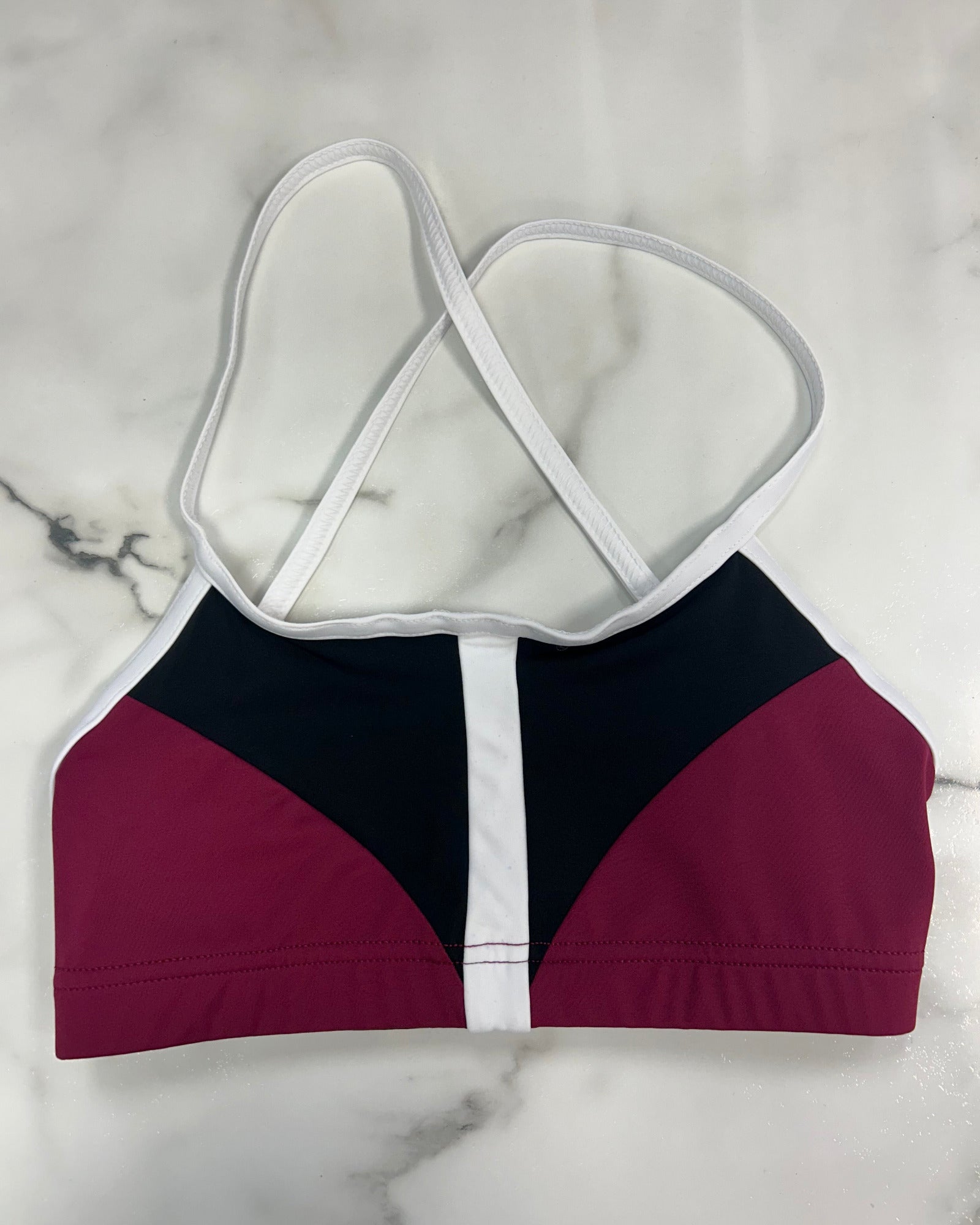 A colorblock dance bra top with a color-blocked design in burgundy, black, and white, featuring thin white crossover shoulder straps, laid flat on a white marble surface.