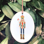 An oval white Christmas ornament featuring a painted nutcracker figure, hanging from a brown ribbon against a background of green holiday leaves and pinecones.