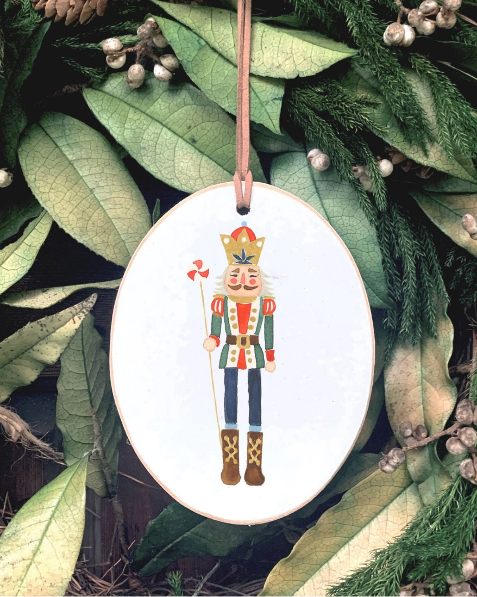 An oval white Christmas ornament featuring a painted nutcracker figure, hanging from a brown ribbon against a background of green holiday leaves and pinecones.