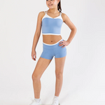 A young girl modeling a light blue dance crop top and matching shorts set with white trim, paired with white sneakers, against a plain white studio background.