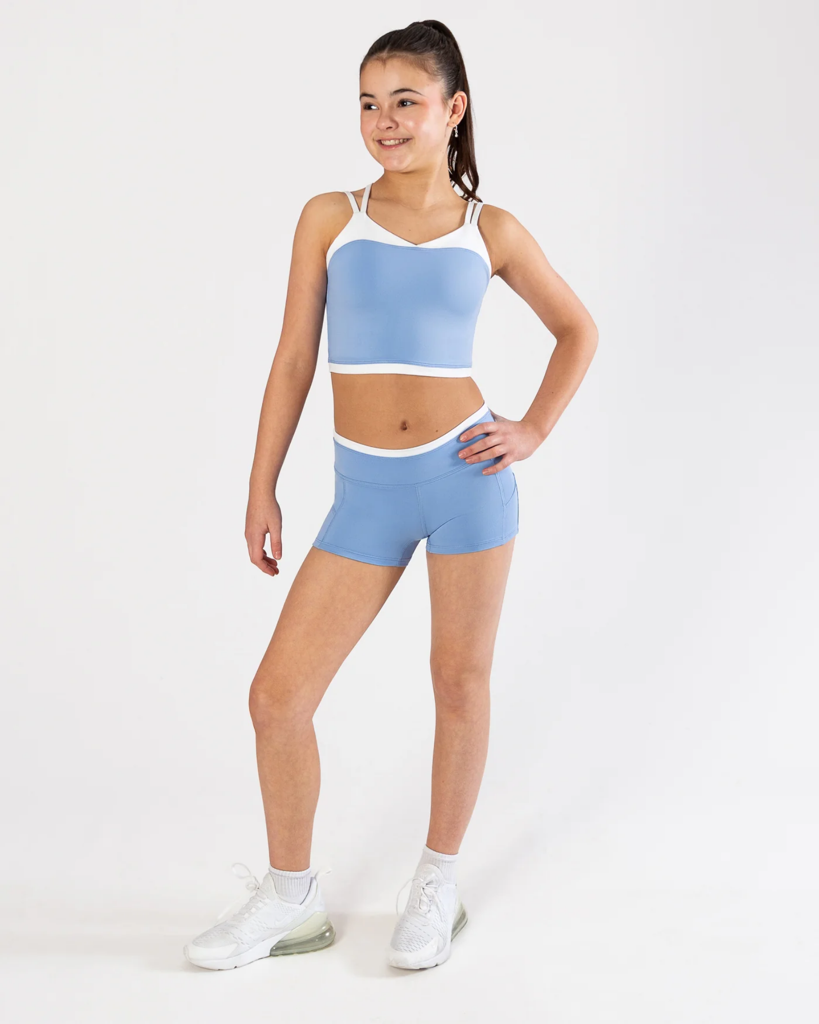 A young girl modeling a light blue dance crop top and matching shorts set with white trim, paired with white sneakers, against a plain white studio background.