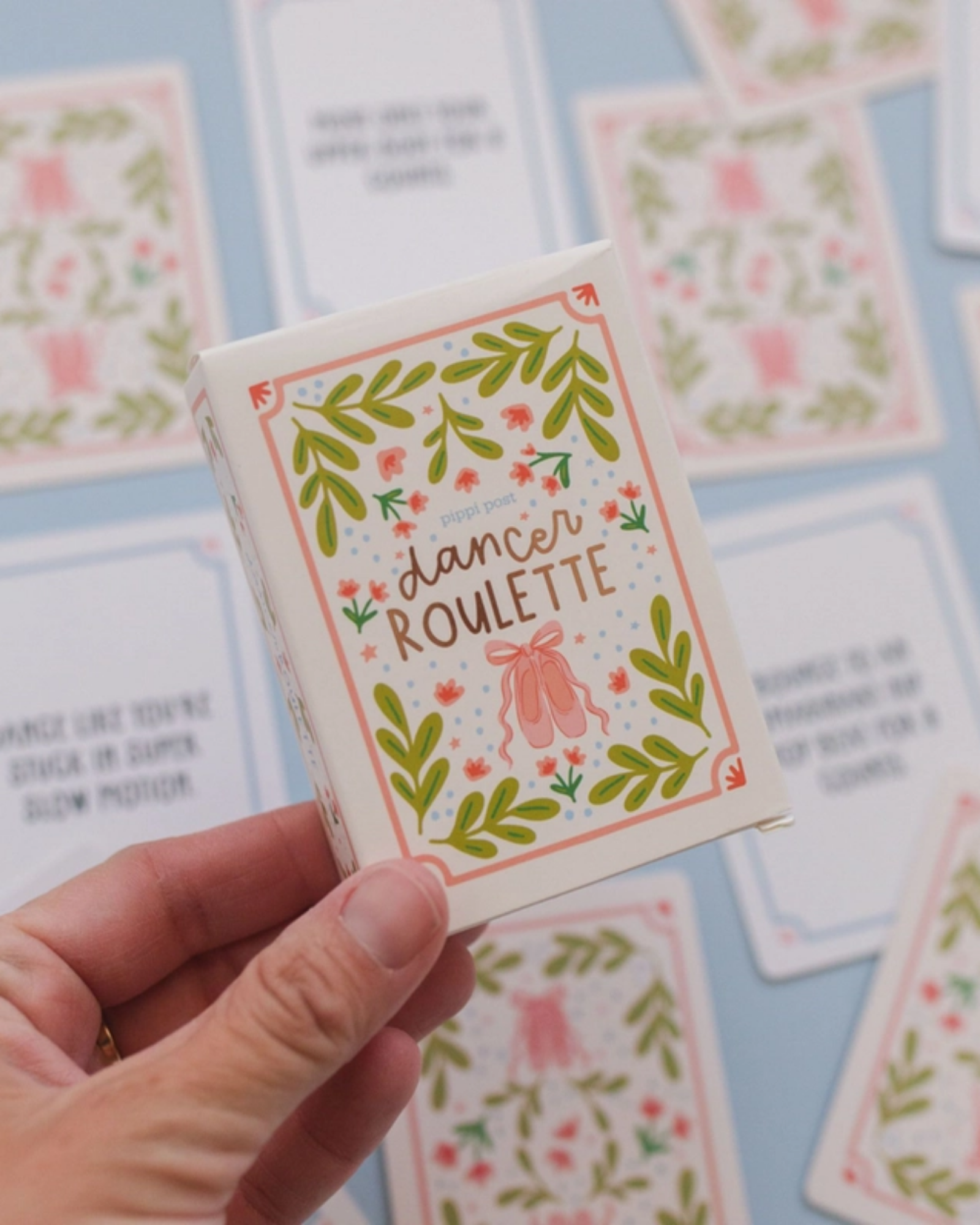 A hand holds a small illustrated card deck box labeled "dancer roulette" with floral borders and pink ballet slippers, against a light blue background with matching cards scattered around.