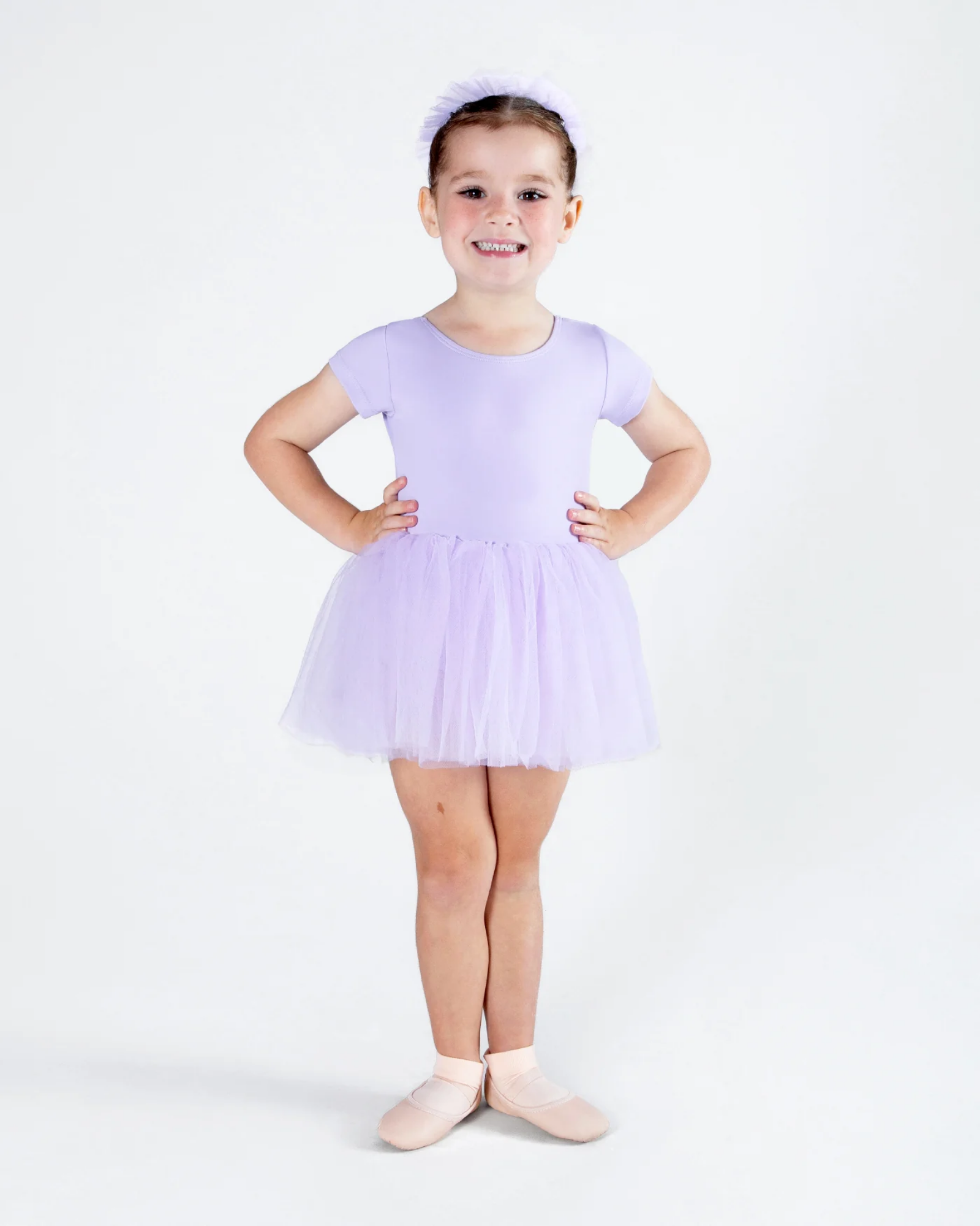 A young girl modeling a light purple ballet leotard with a matching tutu skirt and a matching headband, standing in ballet slippers against a white studio background.
