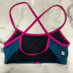 A women's dance bra laid flat on a white marble surface, featuring a color-blocked design with black, teal blue, and bright hot pink trim and straps.
