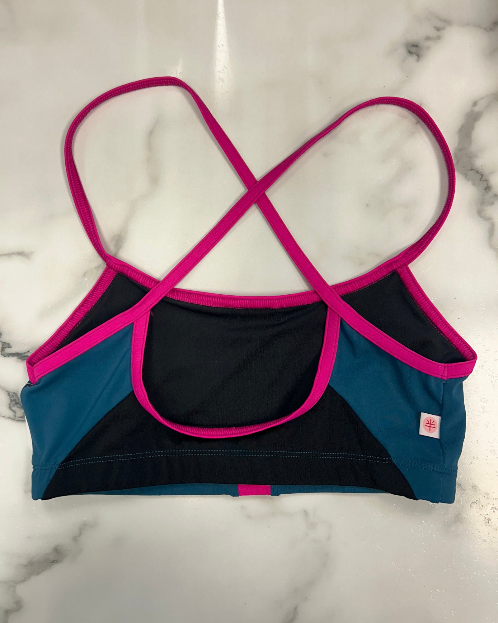 A women's dance bra laid flat on a white marble surface, featuring a color-blocked design with black, teal blue, and bright hot pink trim and straps.