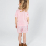 A young girl with blonde pigtails is shown from behind wearing a light pink pajama t-shirt, light pink patterned shorts, and brown boots against a plain white studio background.