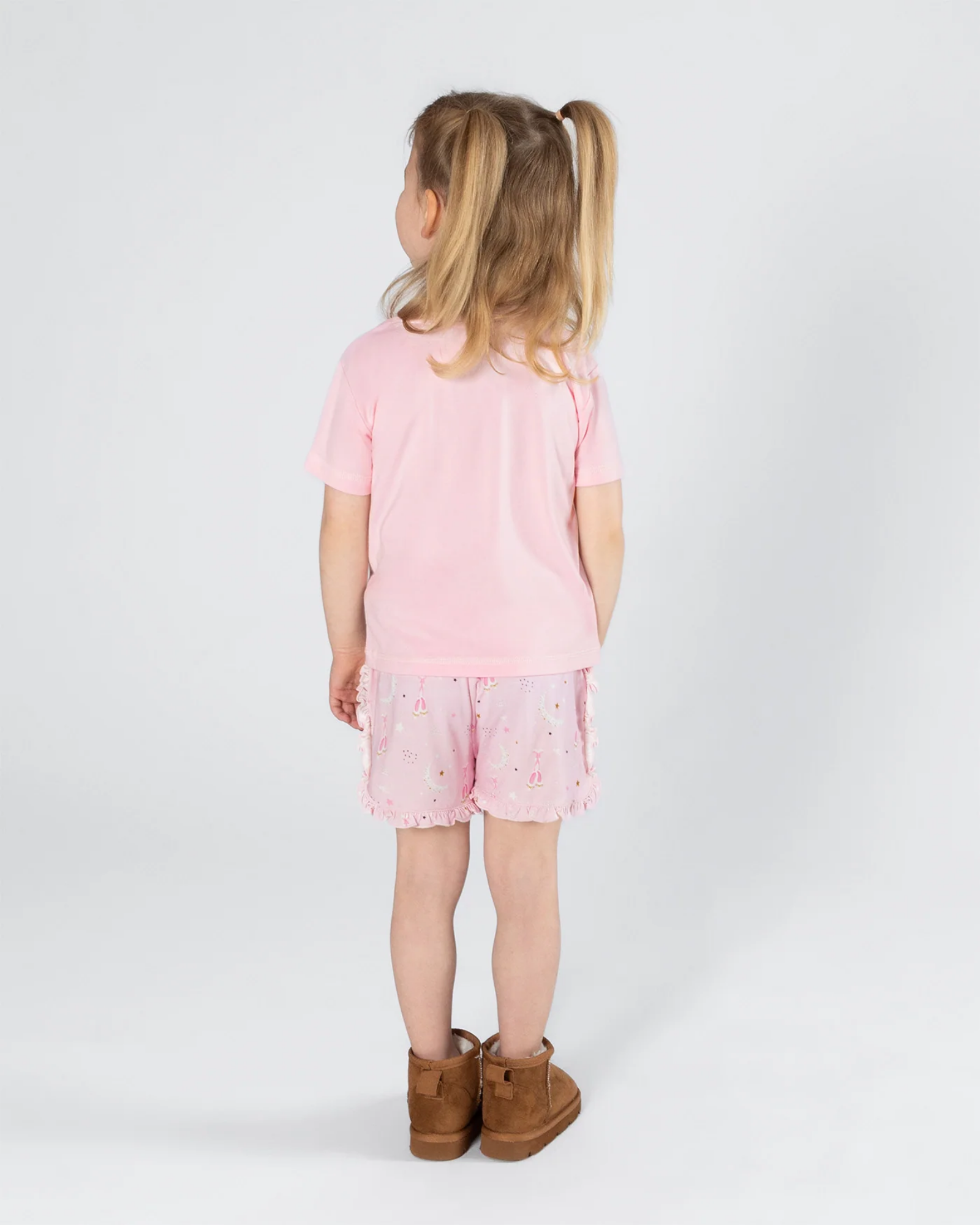 A young girl with blonde pigtails is shown from behind wearing a light pink pajama t-shirt, light pink patterned shorts, and brown boots against a plain white studio background.