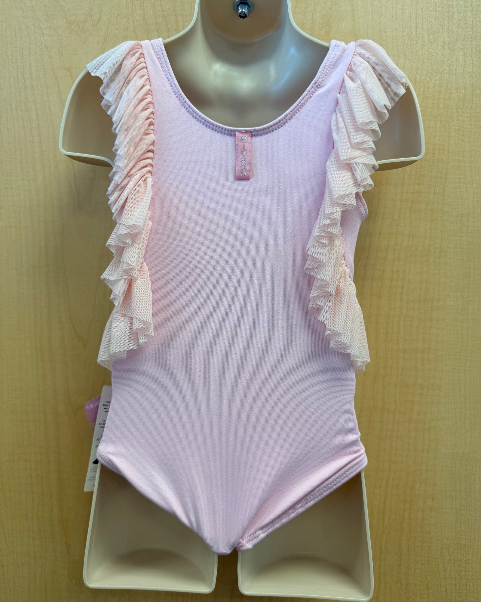 A light pink baby or toddler leotard with ruffled shoulder straps, displayed on a beige mannequin torso against a light wood background.