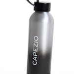 A sleek, metallic gray to black gradient water bottle with a black cap and loop handle, featuring the brand name 'CAPEZIO' printed vertically in white. The bottle is photographed standing on a light beige surface against a plain white background.