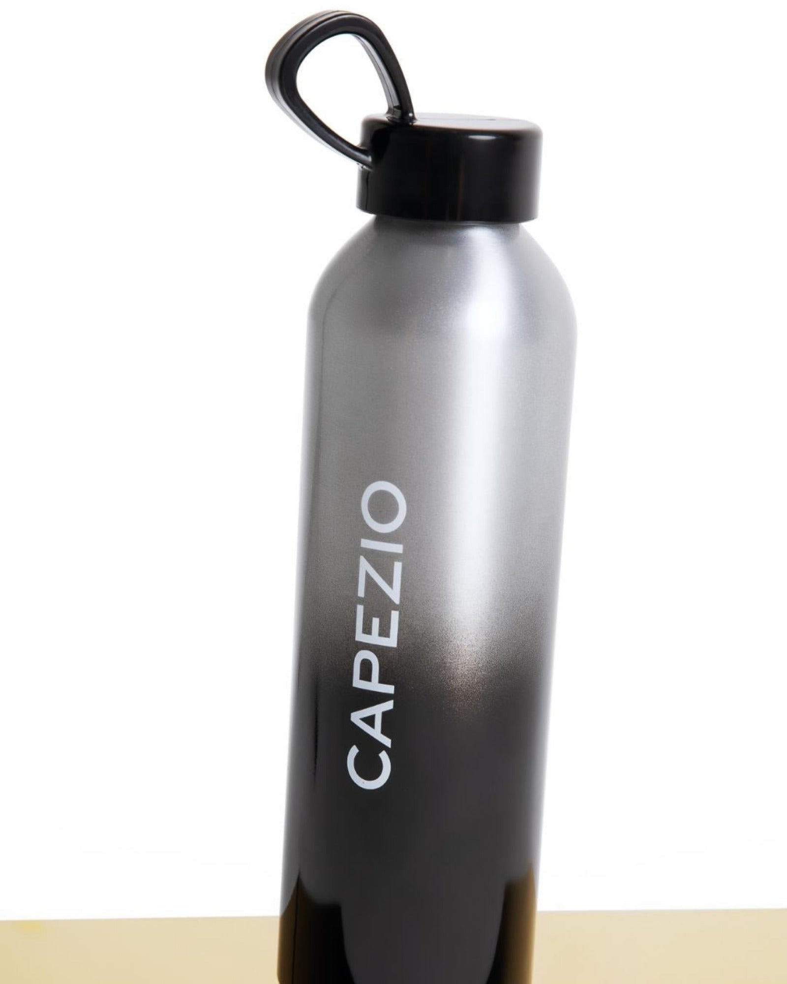 A sleek, metallic gray to black gradient water bottle with a black cap and loop handle, featuring the brand name 'CAPEZIO' printed vertically in white. The bottle is photographed standing on a light beige surface against a plain white background.