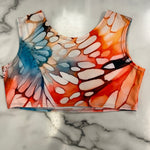 A sleeveless crop top with a colorful butterfly wing pattern in orange, red, blue, and white, laid flat on a white marble surface.