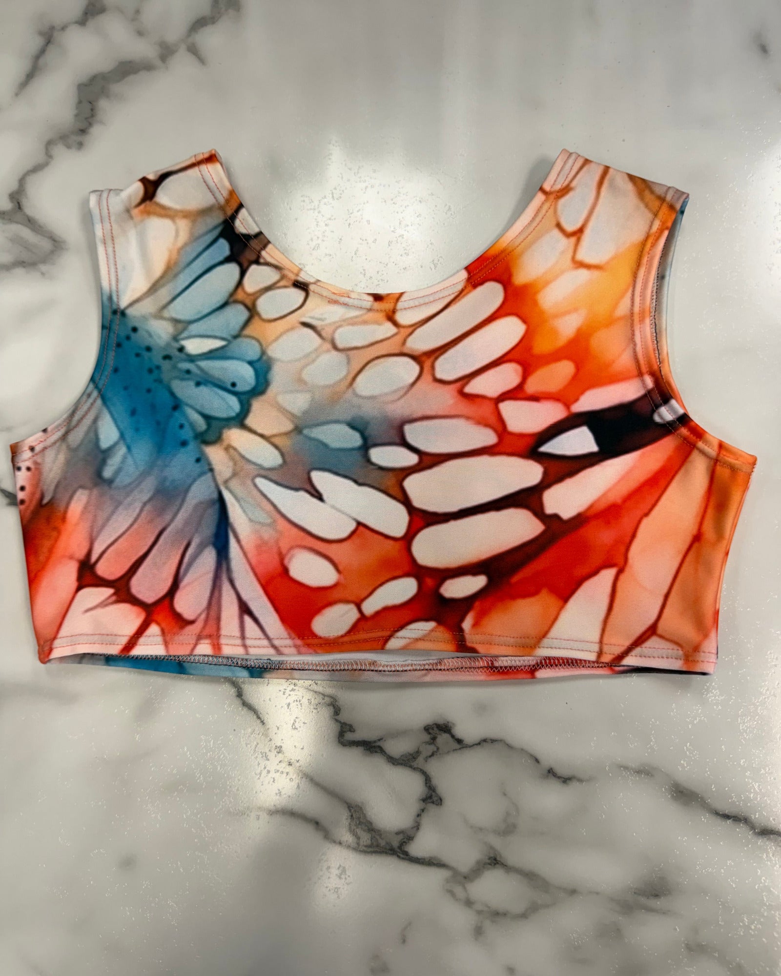 A sleeveless crop top with a colorful butterfly wing pattern in orange, red, blue, and white, laid flat on a white marble surface.