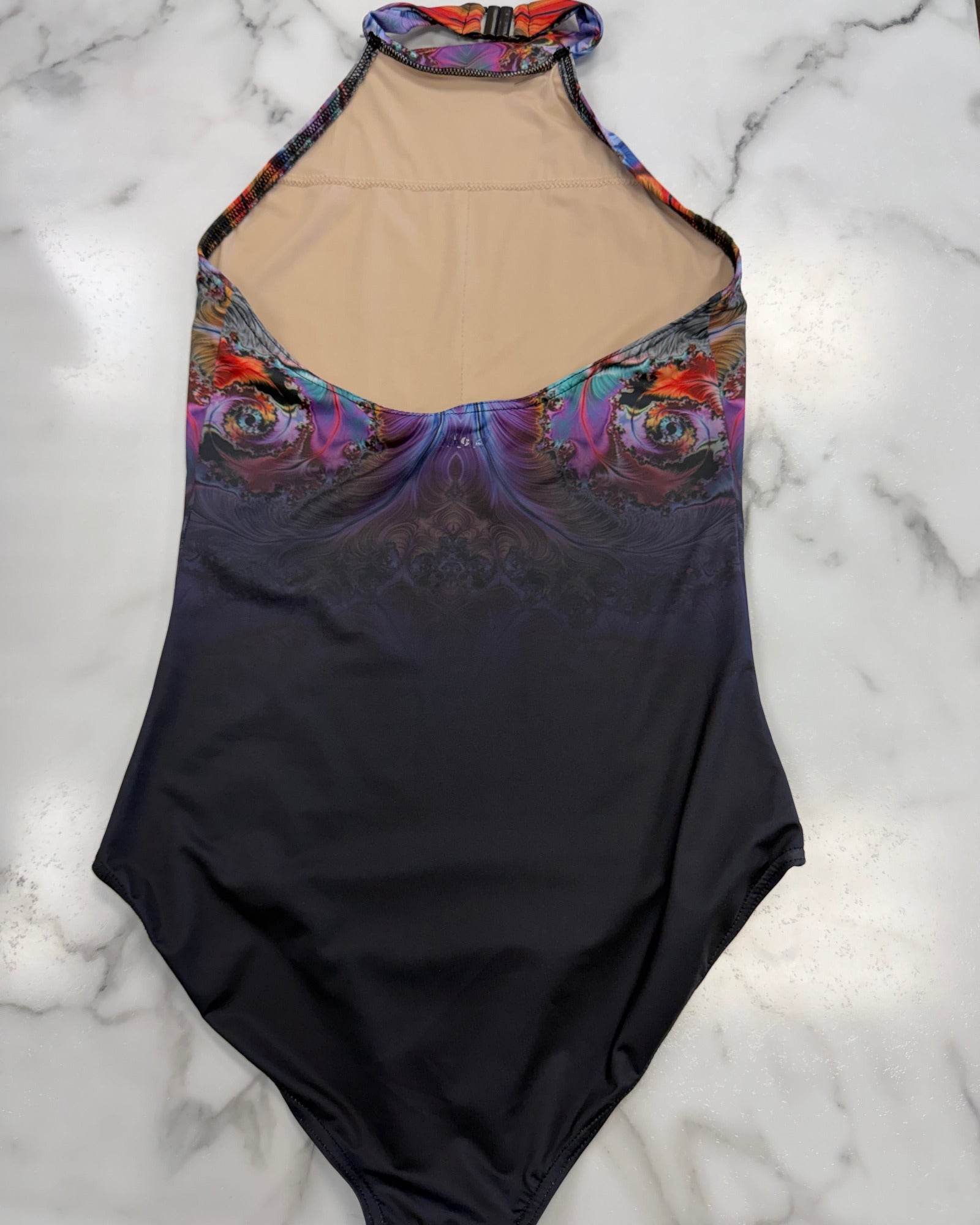 A halter-neck one-piece leotard laid flat on a white marble surface. The leotard features a dark navy-to-black gradient lower body, a colorful abstract floral pattern across the bust and upper waist, and a beige mesh or skin-tone inner lining visible at the neckline.