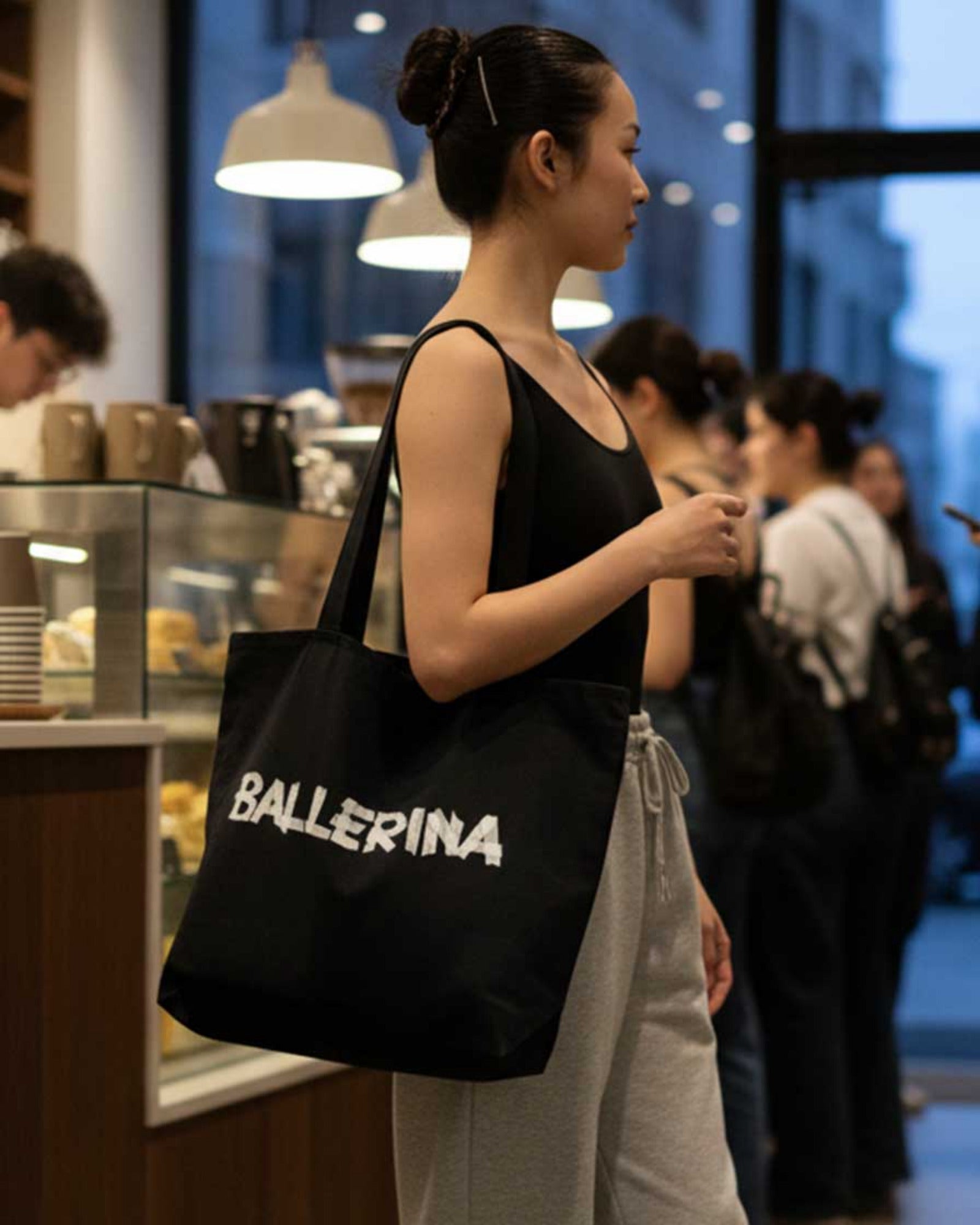 Woman holding a black tote bag with 'BALLERINA' text in a casual setting.