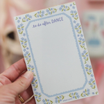 A hand holding a small notepad with a floral border and the words 'to do after DANCE' at the top, against a soft pink blurred background.