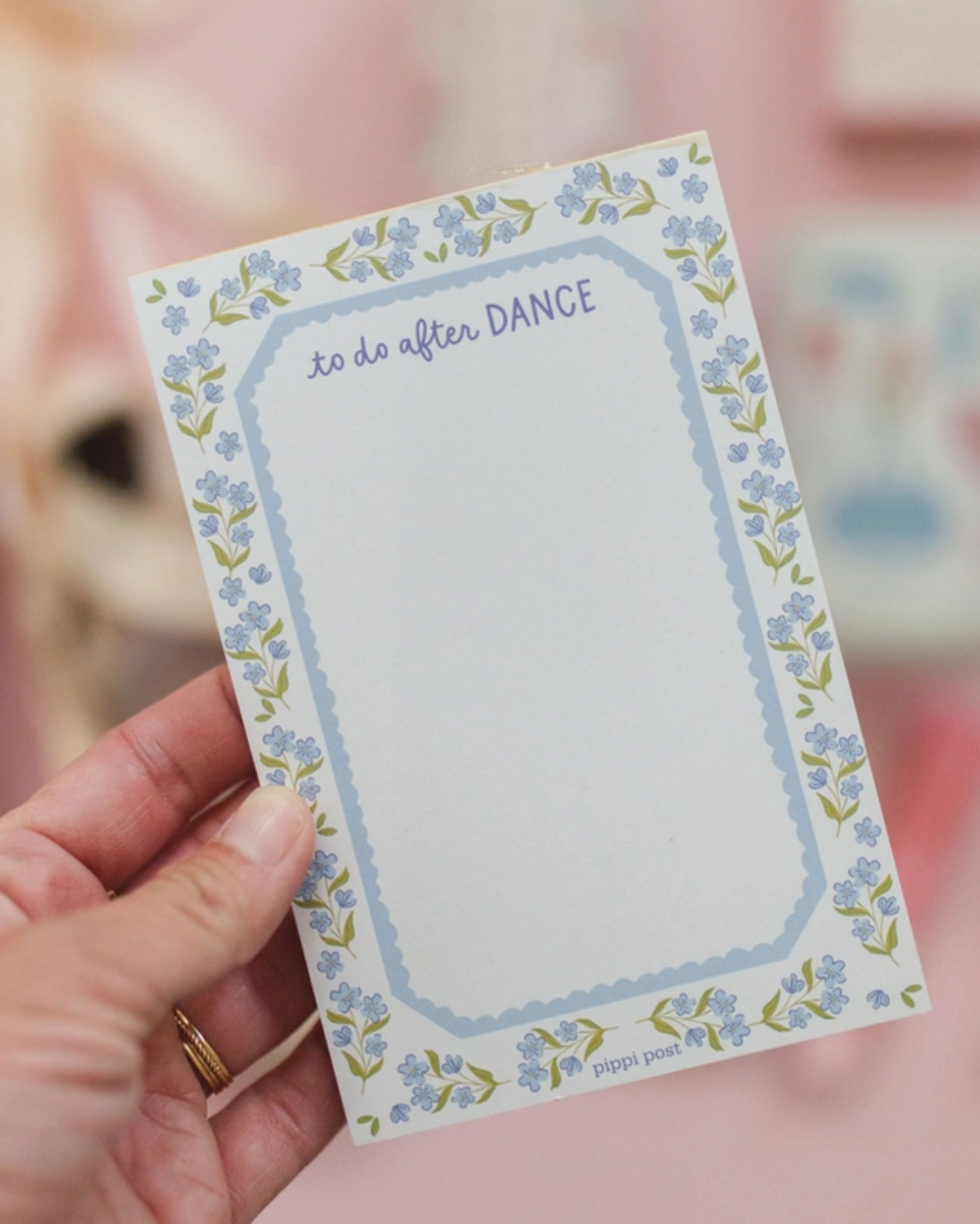 A hand holding a small notepad with a floral border and the words 'to do after DANCE' at the top, against a soft pink blurred background.