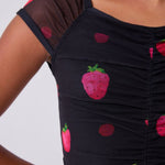 Close-up of a black ruched leotard top with a strawberry and dot print, worn by a model against a light gray background.