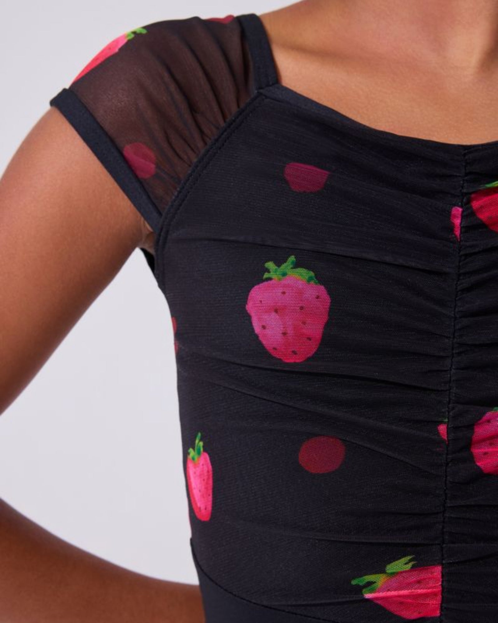 Close-up of a black ruched leotard top with a strawberry and dot print, worn by a model against a light gray background.