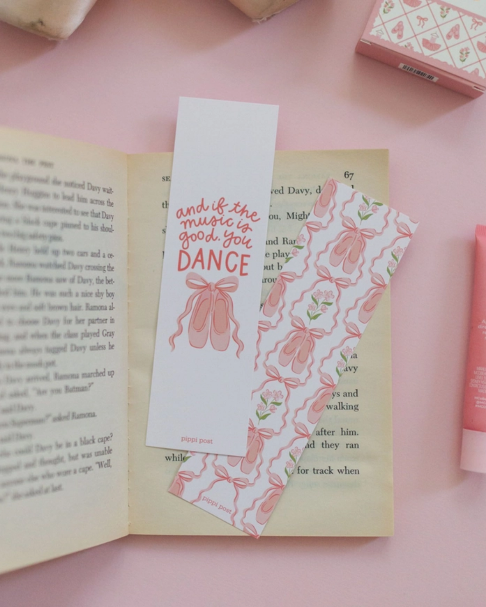 Two decorative paper bookmarks styled on an open book on a pink surface. One bookmark is light pink with pink script text reading 'and the music to make you DANCE' and a pink bow illustration.