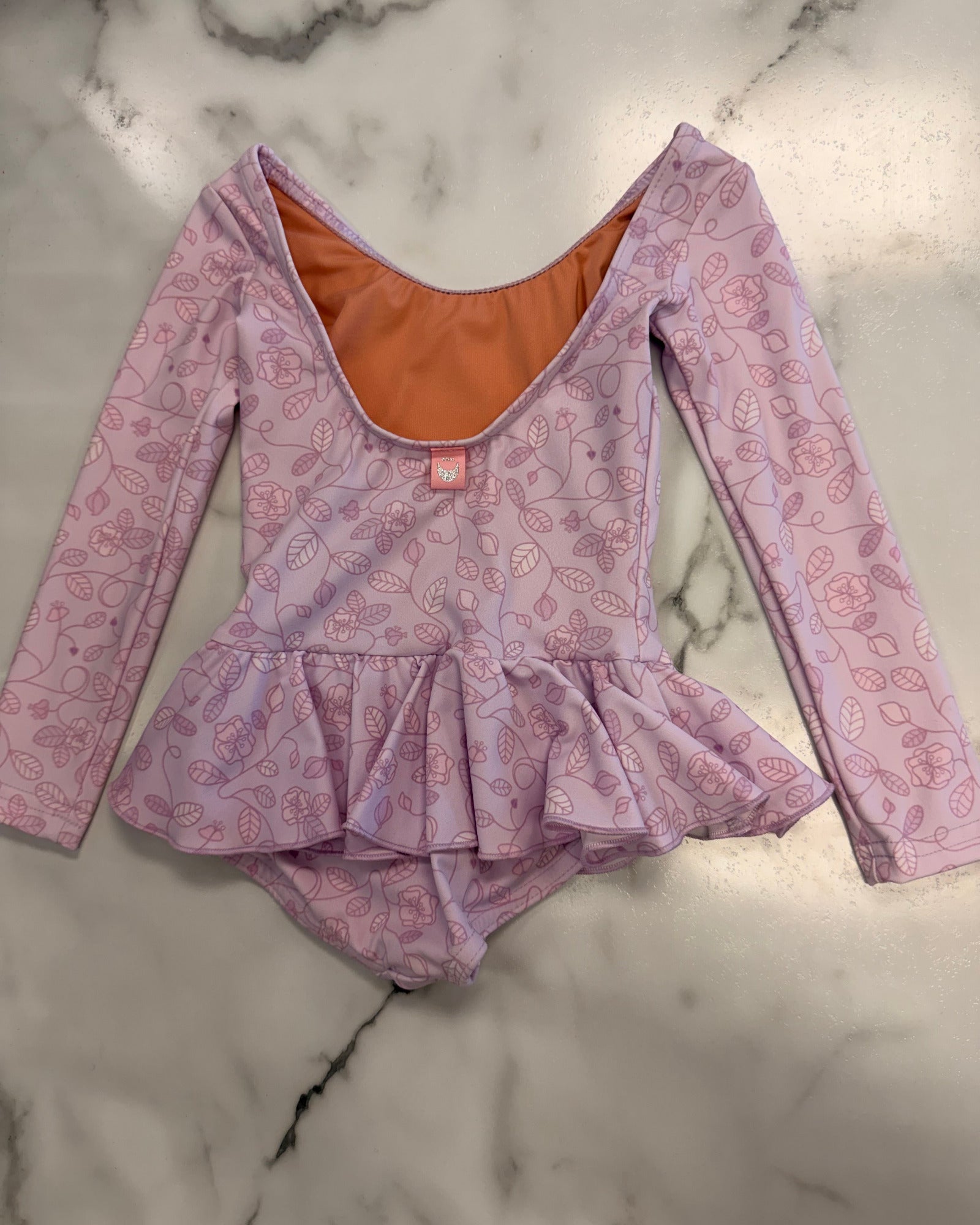 A children's long-sleeve leotard in light purple with a pink floral pattern, featuring a ruffled peplum waist and a contrasting orange inner lining, laid flat on a white marble surface.