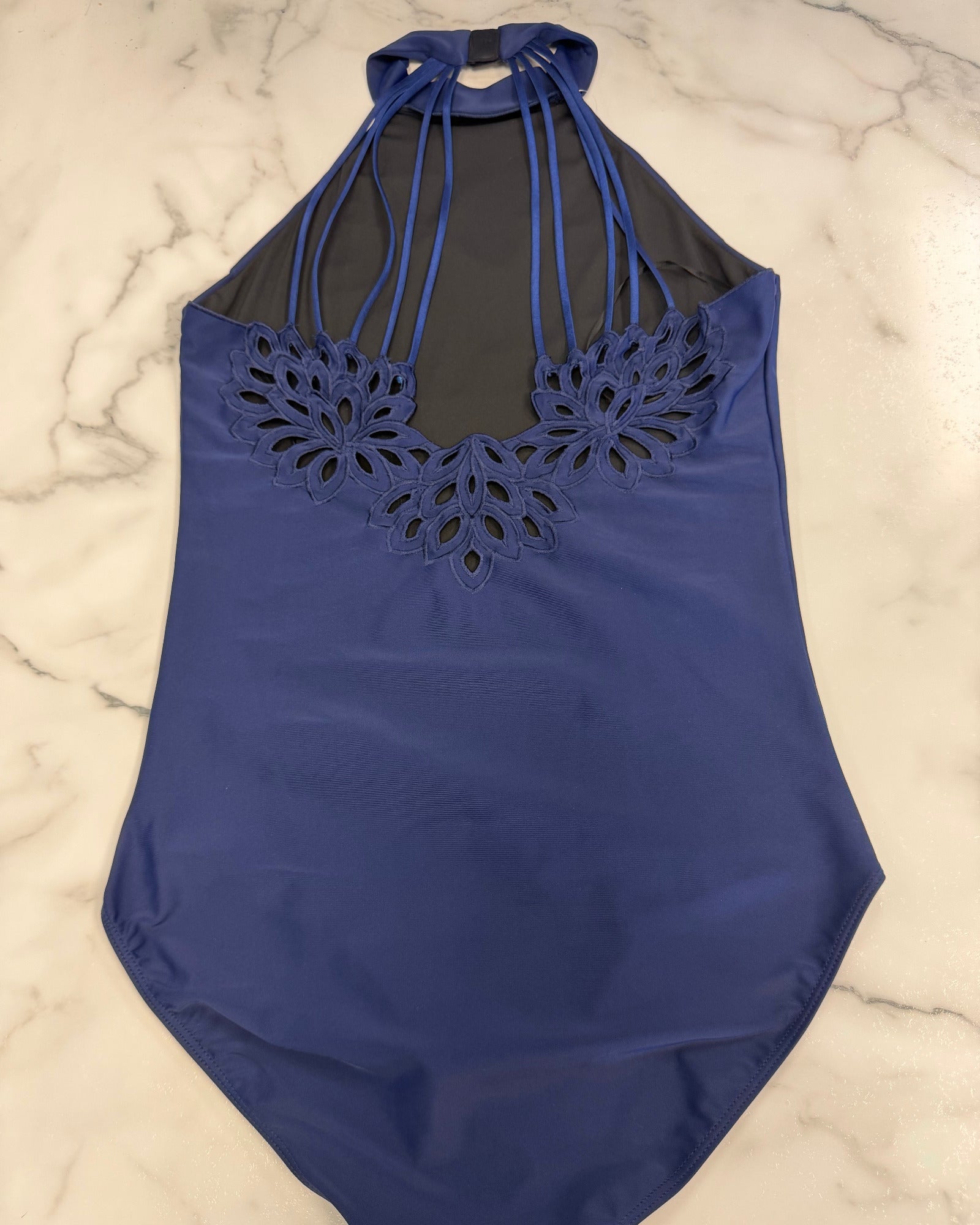 A royal blue halter-neck one-piece leotard with a floral cutout pattern across the chest, laid flat on a white marble surface.