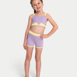 A young girl modeling a lavender and yellow two-piece dance set, consisting of a bra top and matching shorts, against a light gray studio background.