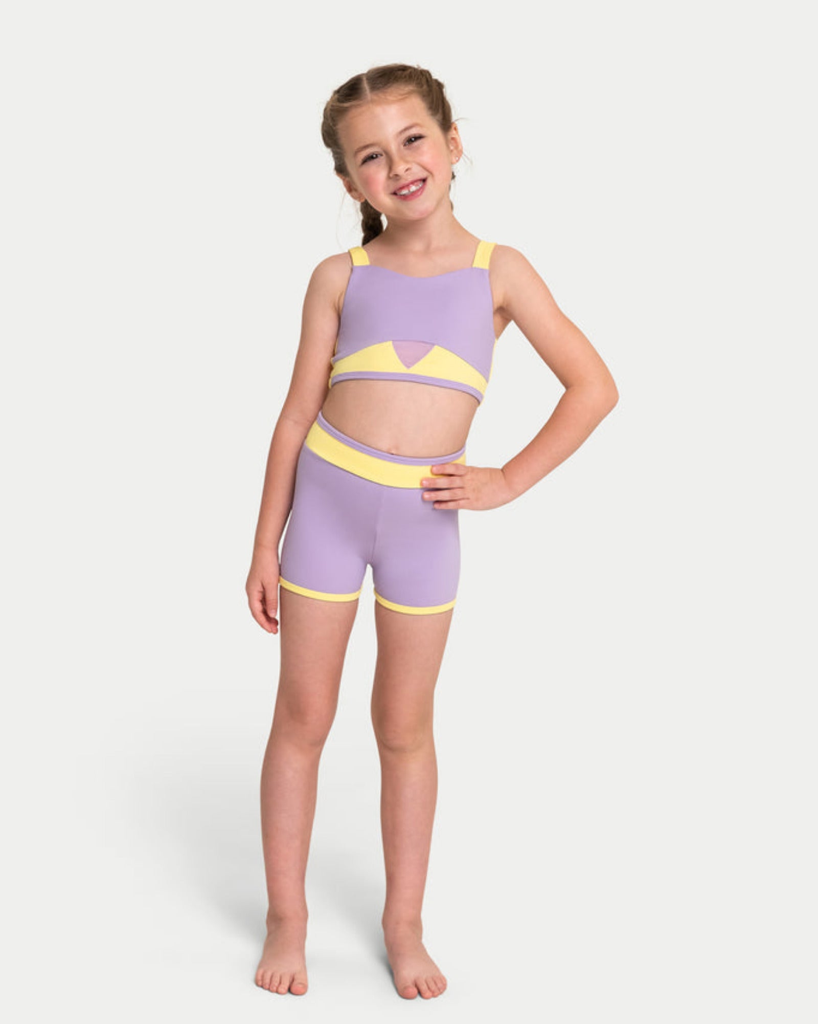 A young girl modeling a lavender and yellow two-piece dance set, consisting of a bra top and matching shorts, against a light gray studio background.