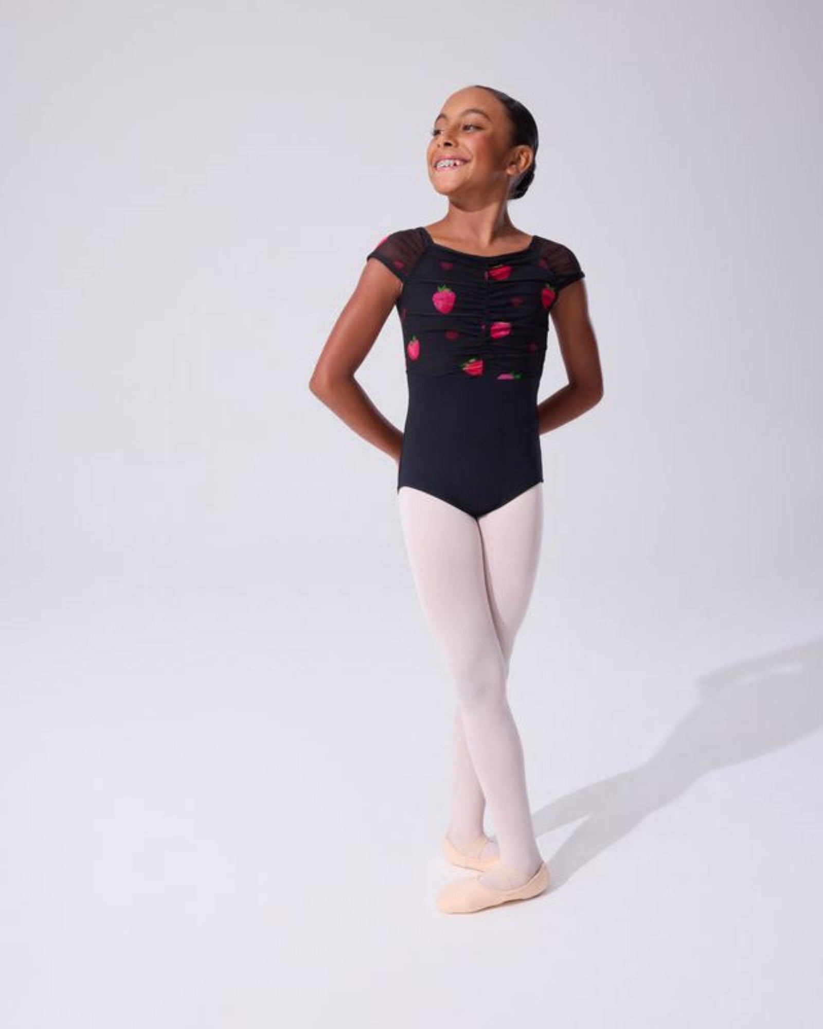 A young girl dancer posing in a black leotard with pink and red floral strawberry patterns, paired with light pink tights and ballet slippers, against a plain white studio background.