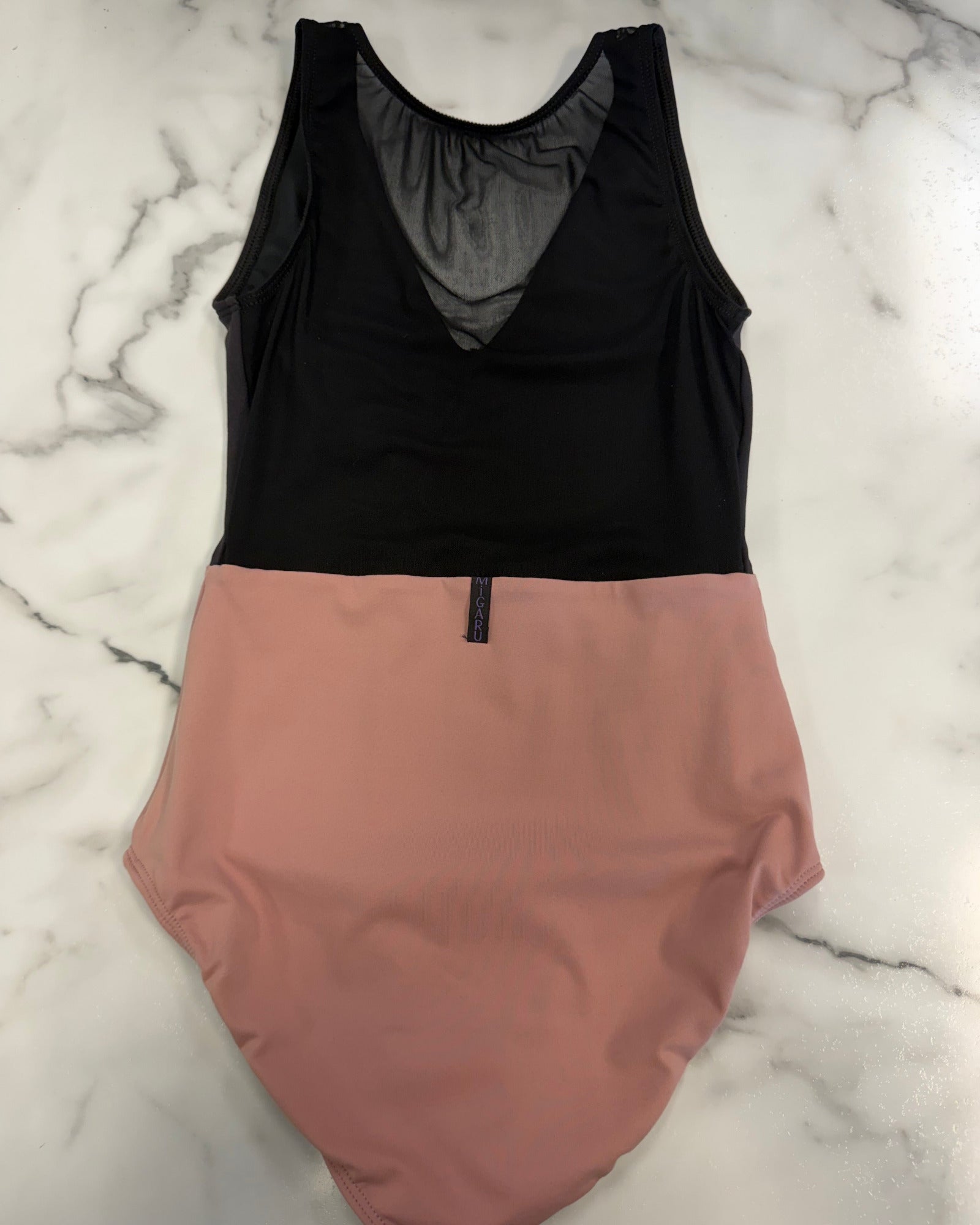 A two-tone sleeveless leotard laid flat on a white marble surface, featuring a black mesh upper section and a dusty pink lower section.