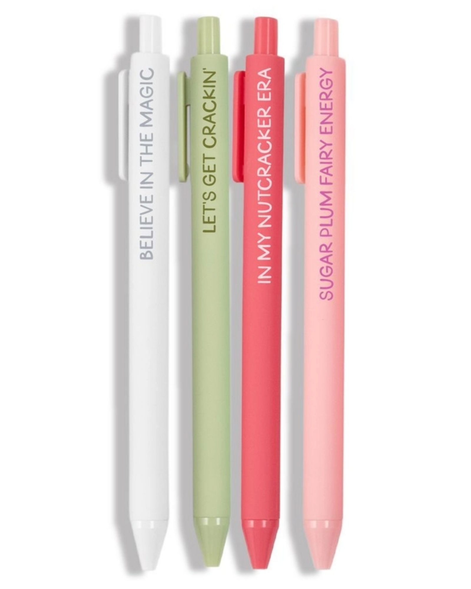 Four slim retractable pens in white, sage green, coral red, and light pink, standing upright on a white background. Each pen has a matching cap and vertical text: 'BELIEVE IN THE MAGIC,' 'LET'S GET CRACKER,' 'IN MY NUTCRACKER ERA,' and 'SUGAR PLUM FAIRY ENERGY.