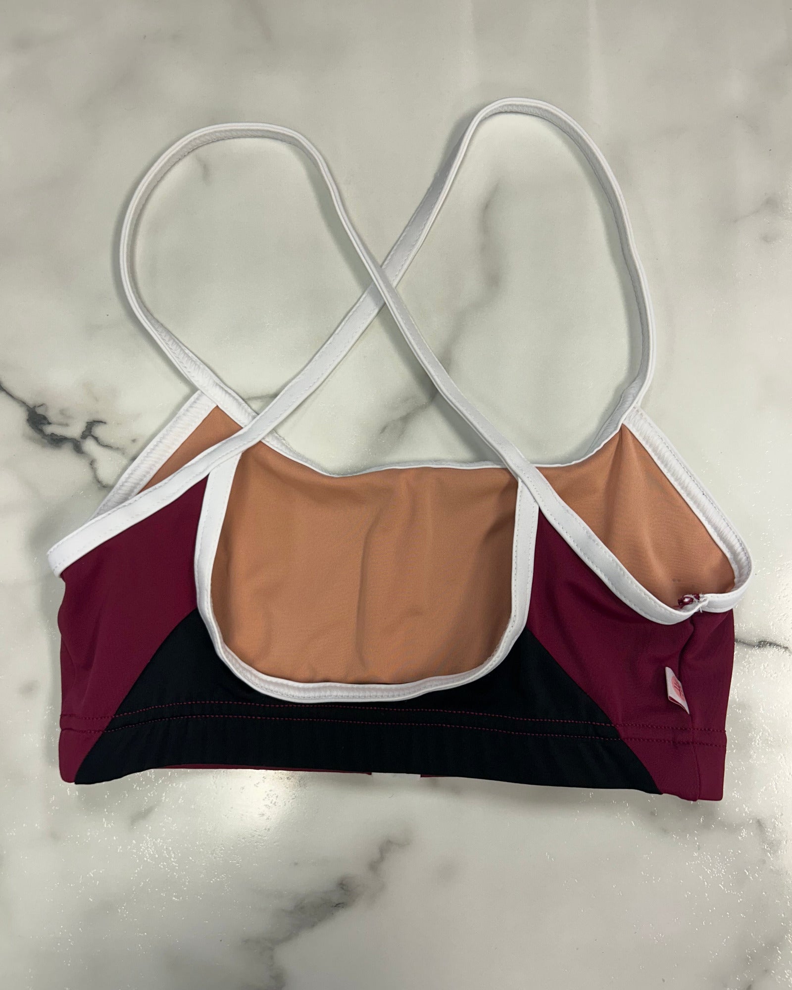 A color-blocked dance bra laid flat on a white marble surface, featuring a nude lining, burgundy side panels, a black bottom panel, and white straps with white trim.