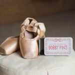 A small rectangular tin labeled 'emergency BOBBY PINS' with a floral border sits beside a pair of satin pointe shoes on a light fabric surface.