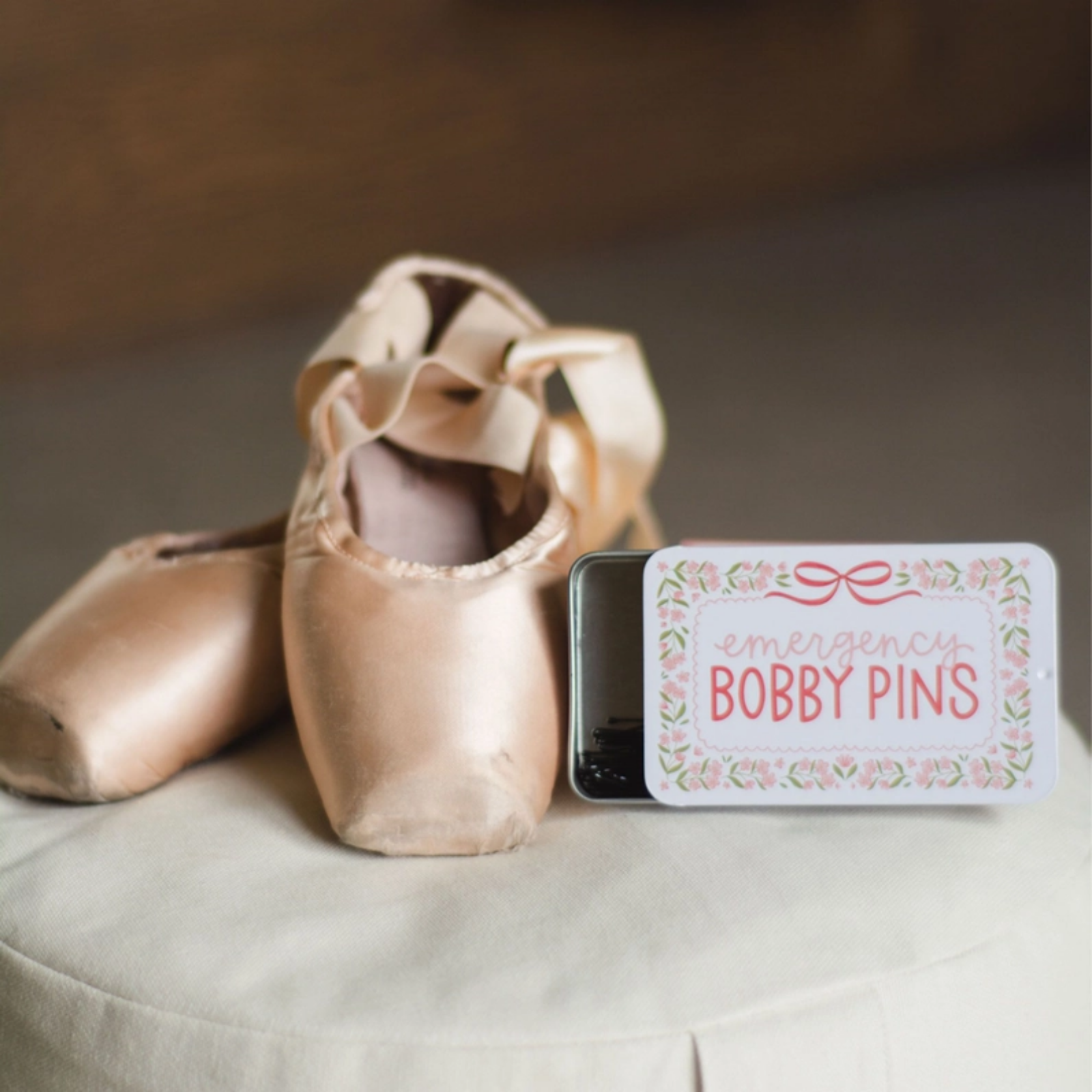 A small rectangular tin labeled 'emergency BOBBY PINS' with a floral border sits beside a pair of satin pointe shoes on a light fabric surface.