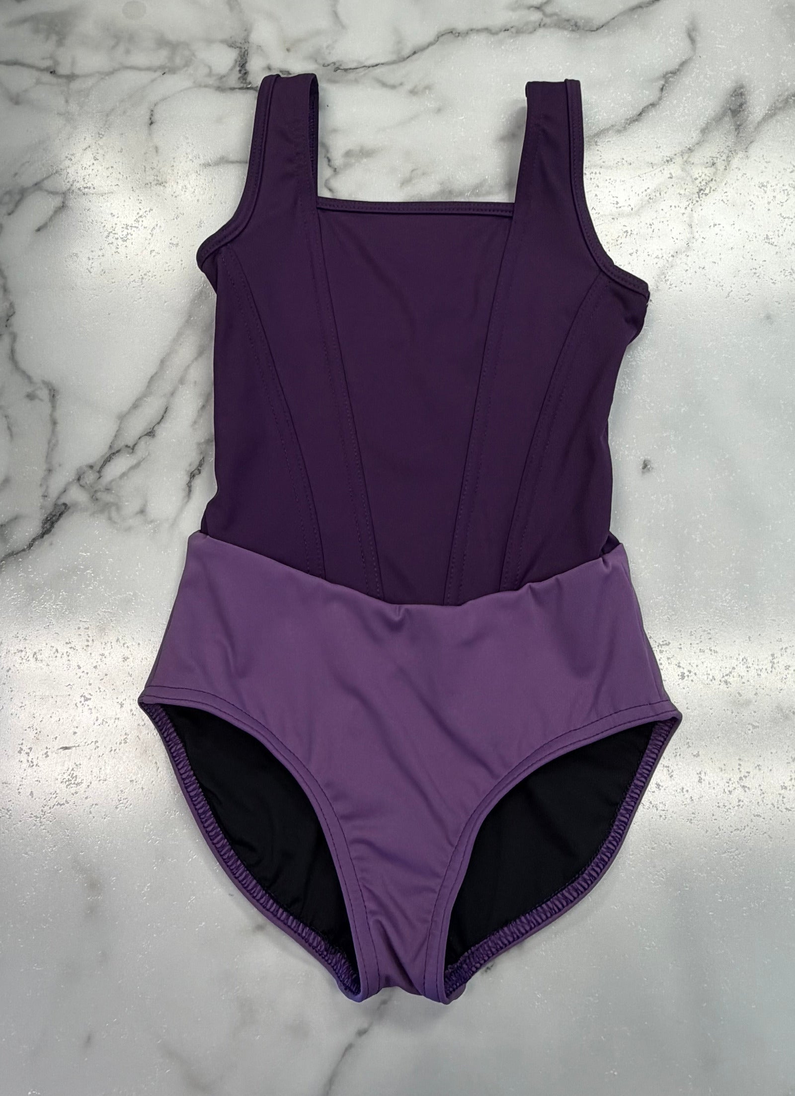 A purple leotard laid flat on a white marble surface, featuring a darker purple bodice and a lighter purple high-waisted bottom with a black lining visible at the legs.