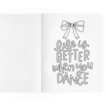 A white open notebook or journal spread on a white background, with black hand-lettered text on the right page reading 'Life is BETTER when you DANCE' beneath a simple bow illustration.