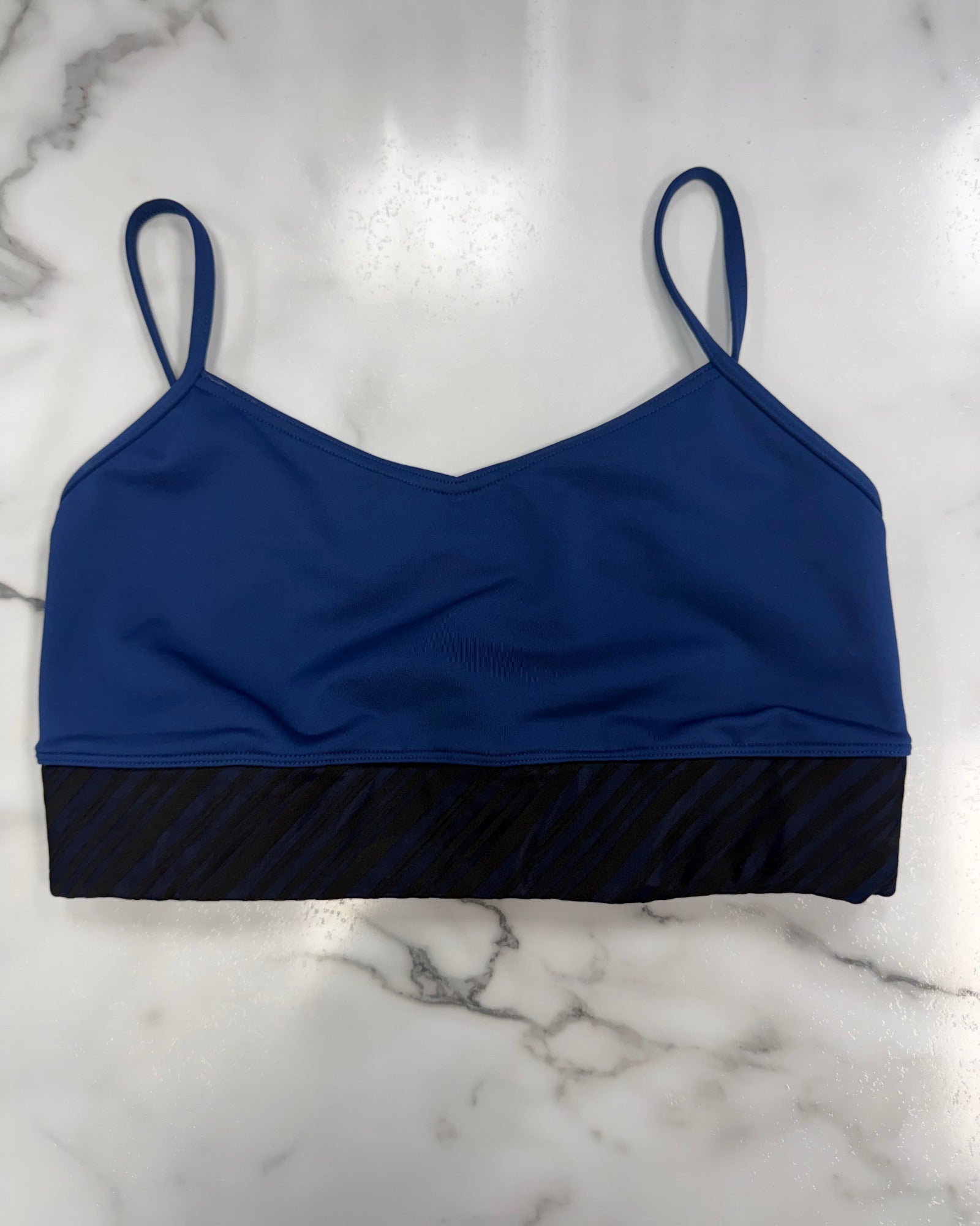 A royal blue dance bra with thin shoulder straps and a black textured horizontal band at the underbust, laid flat on a white marble surface.