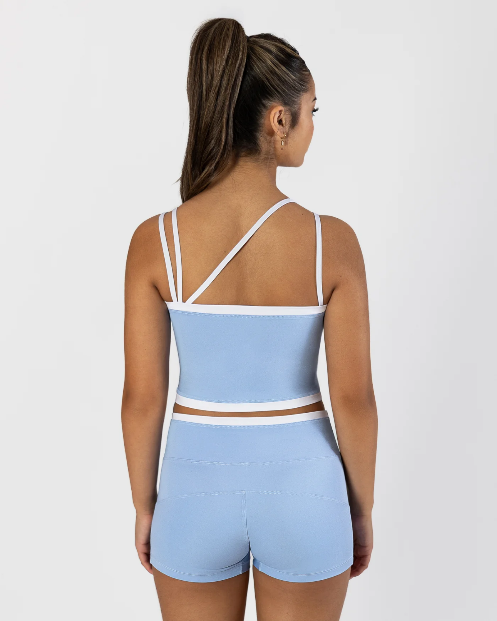 A woman shown from the back wearing a light blue two-piece dancewear set with white trim, consisting of a cropped tank top with crisscross straps and matching high-waisted shorts, against a light gray studio background.
