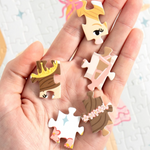 A hand holds several small, illustrated jigsaw puzzle-piece charms with cartoon character faces and hair details, set against a soft-focus background of a completed puzzle.