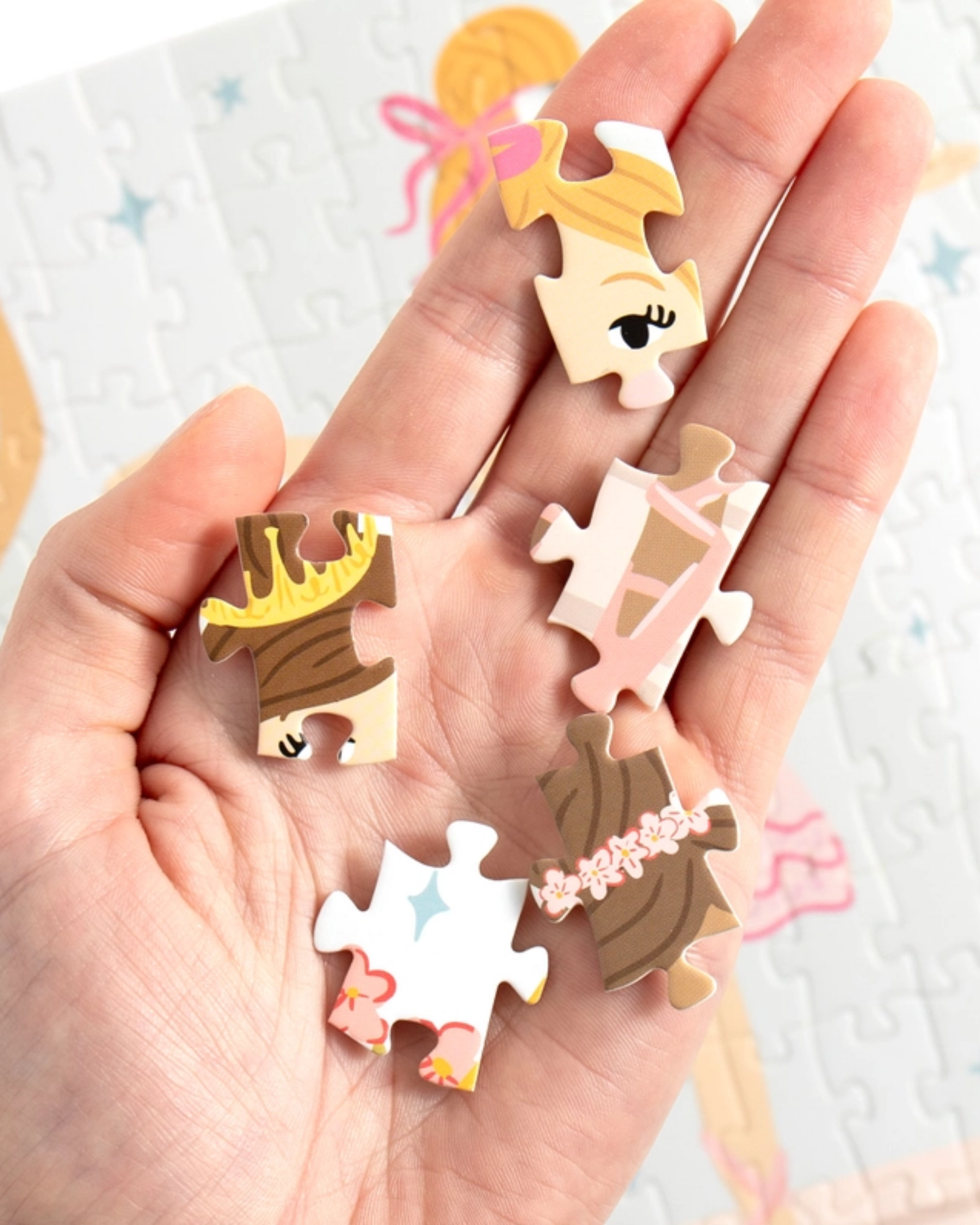 A hand holds several small, illustrated jigsaw puzzle-piece charms with cartoon character faces and hair details, set against a soft-focus background of a completed puzzle.
