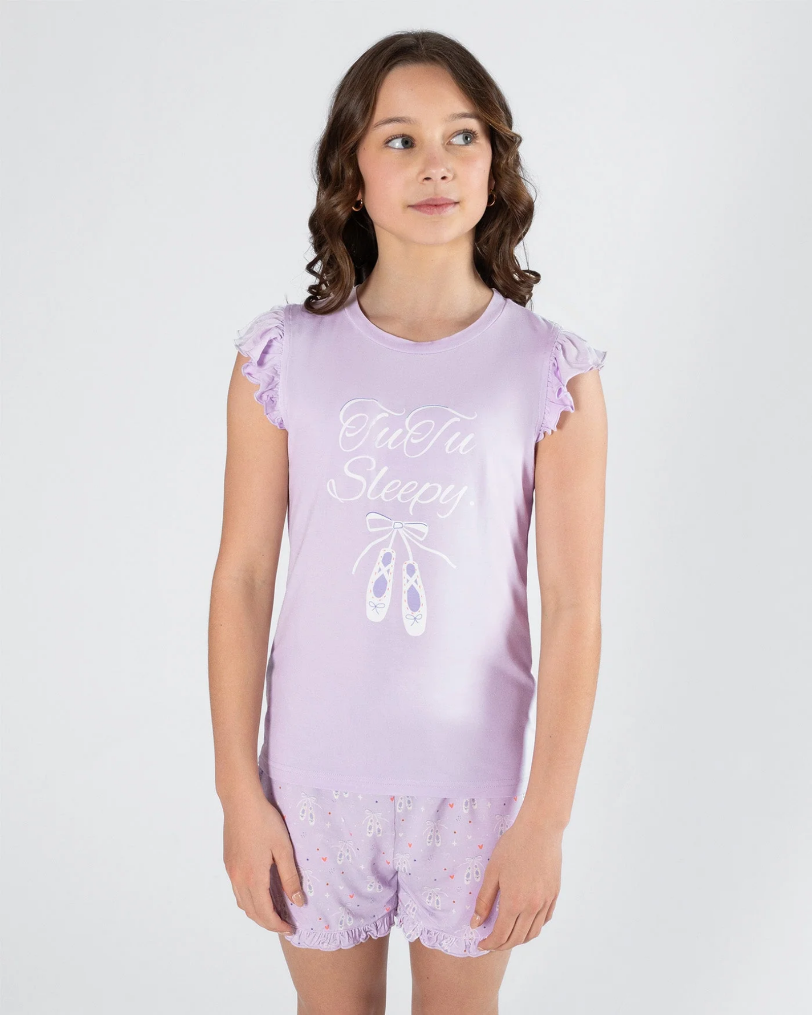 A young girl modeling a lavender pajama set with a ruffled sleeveless top and matching shorts. The top features a white graphic print that reads 'Tutu Sleepy' with a ballet shoe illustration.